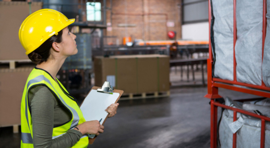 Warehouse worker in a safety helmet inspecting storage racks with a clipboard.