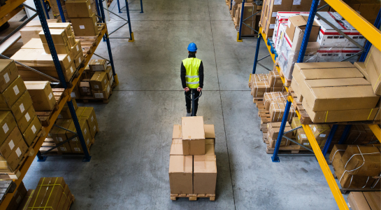 Warehouse worker moving stacked boxes with a pallet jack in a storage aisle.