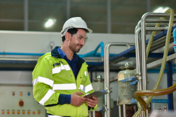 Industrial technician inspecting equipment and taking notes on a clipboard in a factory.