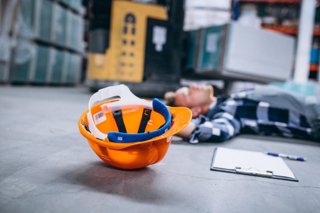 Construction worker lying injured on the floor near a safety helmet in a warehouse.