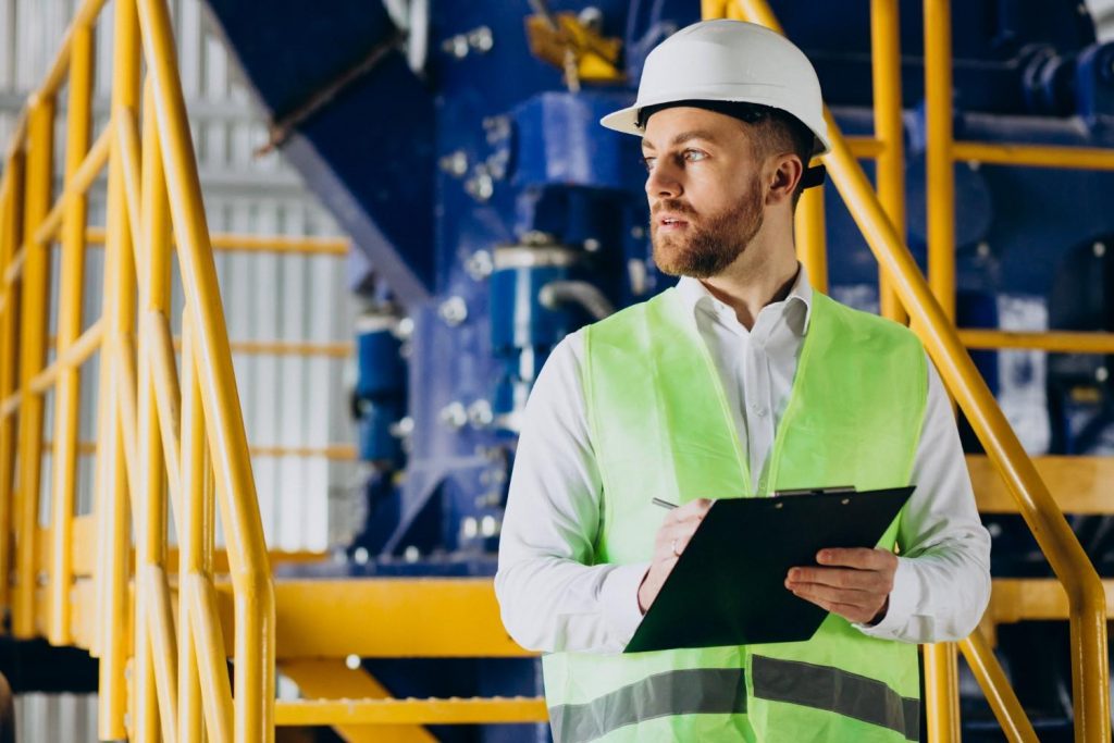 Industrial worker wearing a hard hat and safety vest inspecting equipment with a clipboard.